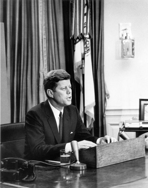 Fotografie alb-negru în care președintele John F. Kennedy este surprins din lateral, așezat la biroul său din Biroul Oval. Citește de pe niște foi în fața mai multor microfoane de emisie, într-un cadru de studio cu echipamente tehnice parțial vizibile în stânga.