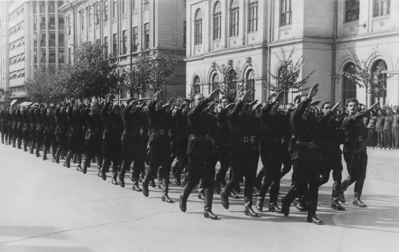 O fotografie istorică alb-negru ce surprinde o coloană masivă de bărbați aparținând Mișcării Legionare, care defilează pe o stradă largă din București. Aceștia poartă cămăși verzi de culoare închisă, pantaloni și cizme lungi de tip militar. Toți bărbații din rândurile din față au brațul drept ridicat în sus, executând salutul fascist (roman). Pe fundal se disting clădiri urbane masive cu arhitectură clasică interbelică.
